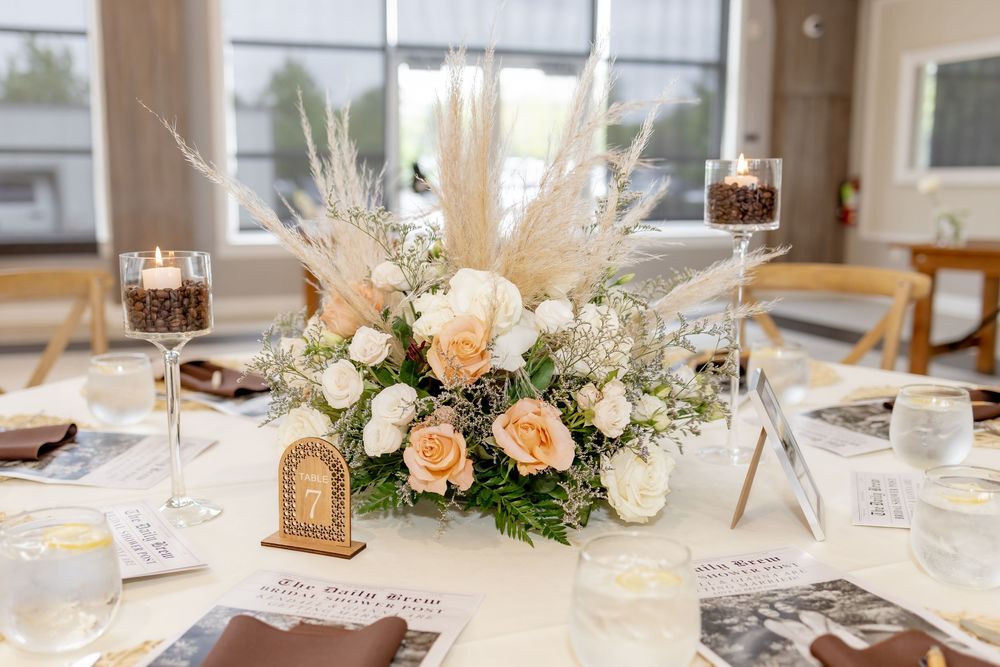 Elegant wedding table centerpiece with peach roses, white flowers, pampas grass, and candles in coffee beans on white linen.