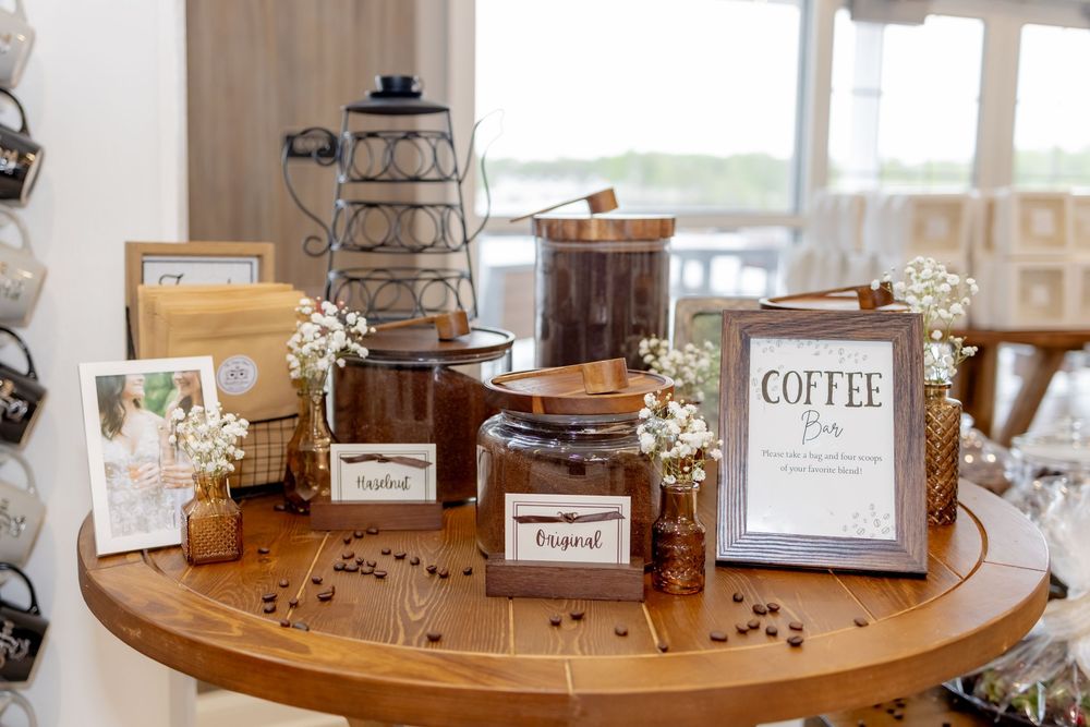 Rustic coffee bar display on round wooden table with labeled jars, coffee beans, white flowers, and a framed Coffee Bar sign.