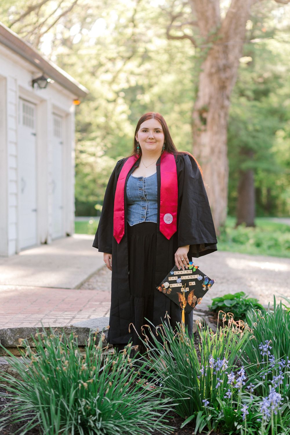 Young woman in black graduation gown with red stole holds decorated cap outdoors among green gardens.