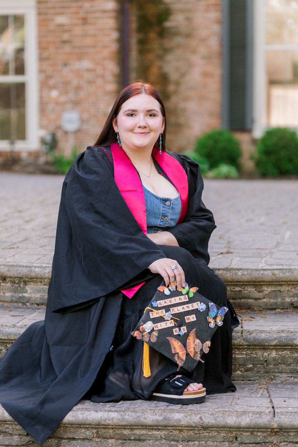 Young woman in black and pink graduation gown sits on stone steps holding decorated cap outdoors.