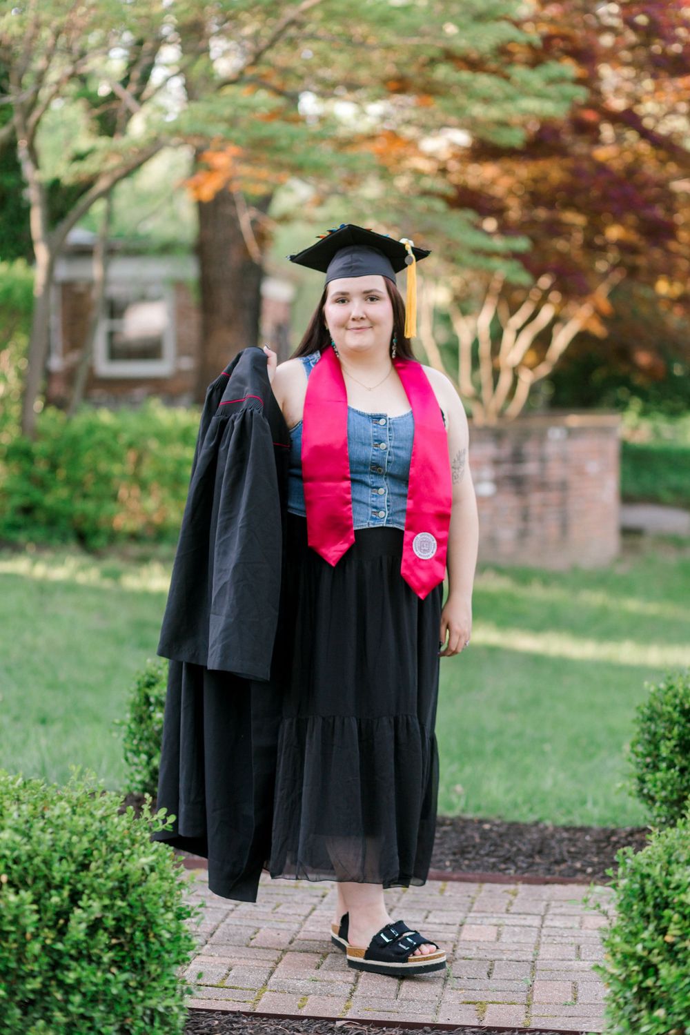 Smiling graduate in cap and gown holding black robe with red stole on brick path surrounded by green bushes.