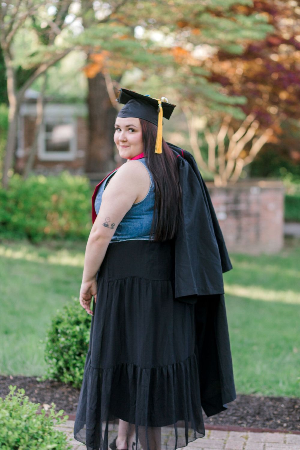 Graduate in black cap and gown smiles over shoulder outdoors, wearing black tulle skirt with yellow tassel.