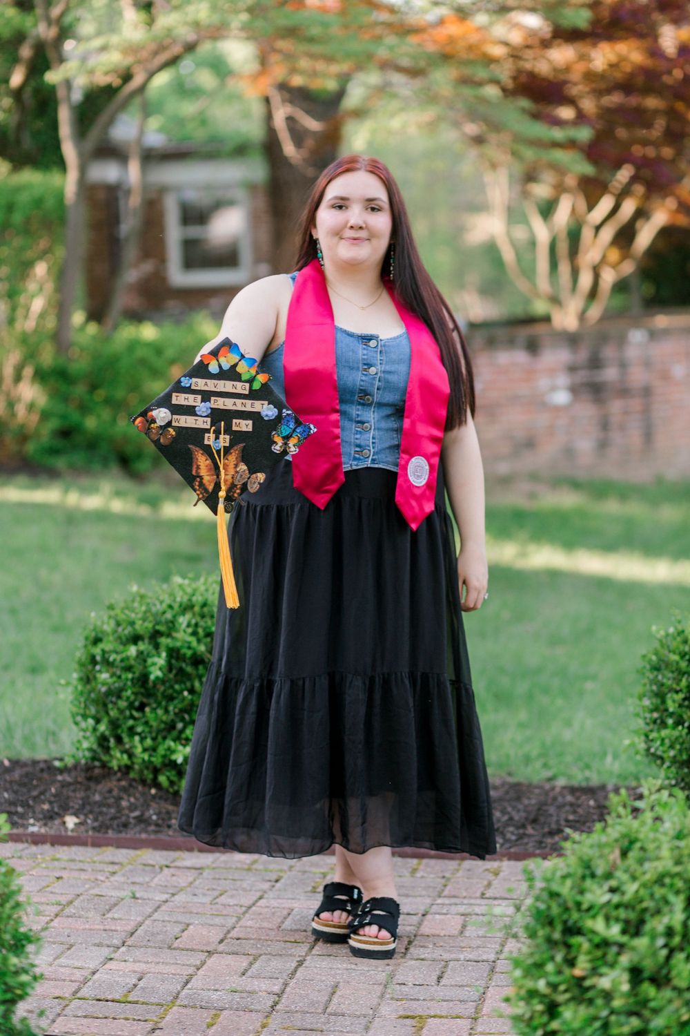Young woman in graduation attire holding decorated cap, wearing pink stole and black skirt on brick path outdoors.