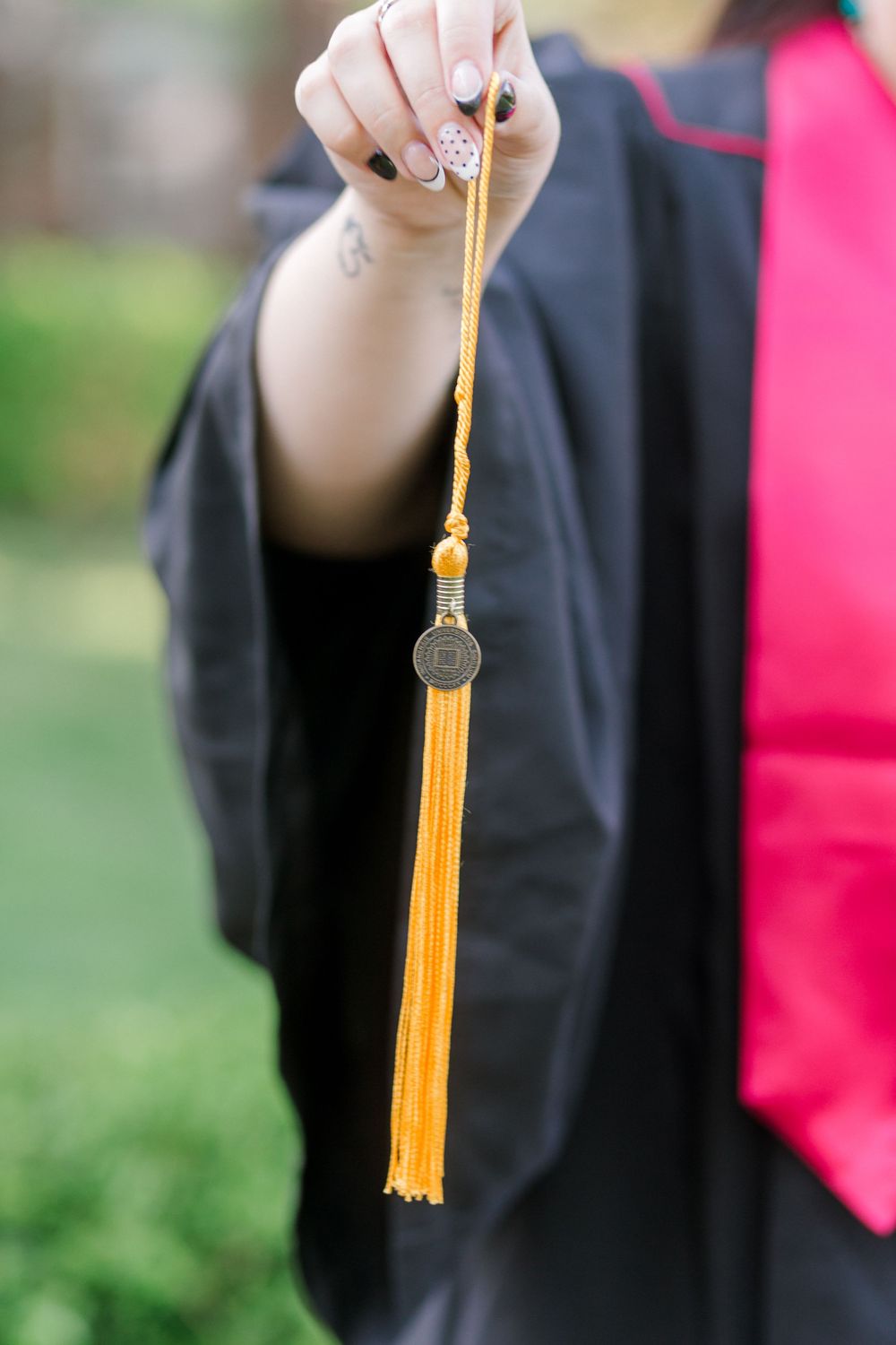 A graduate holds up a gold tassel with a medallion charm, wearing a black graduation gown with red trim.