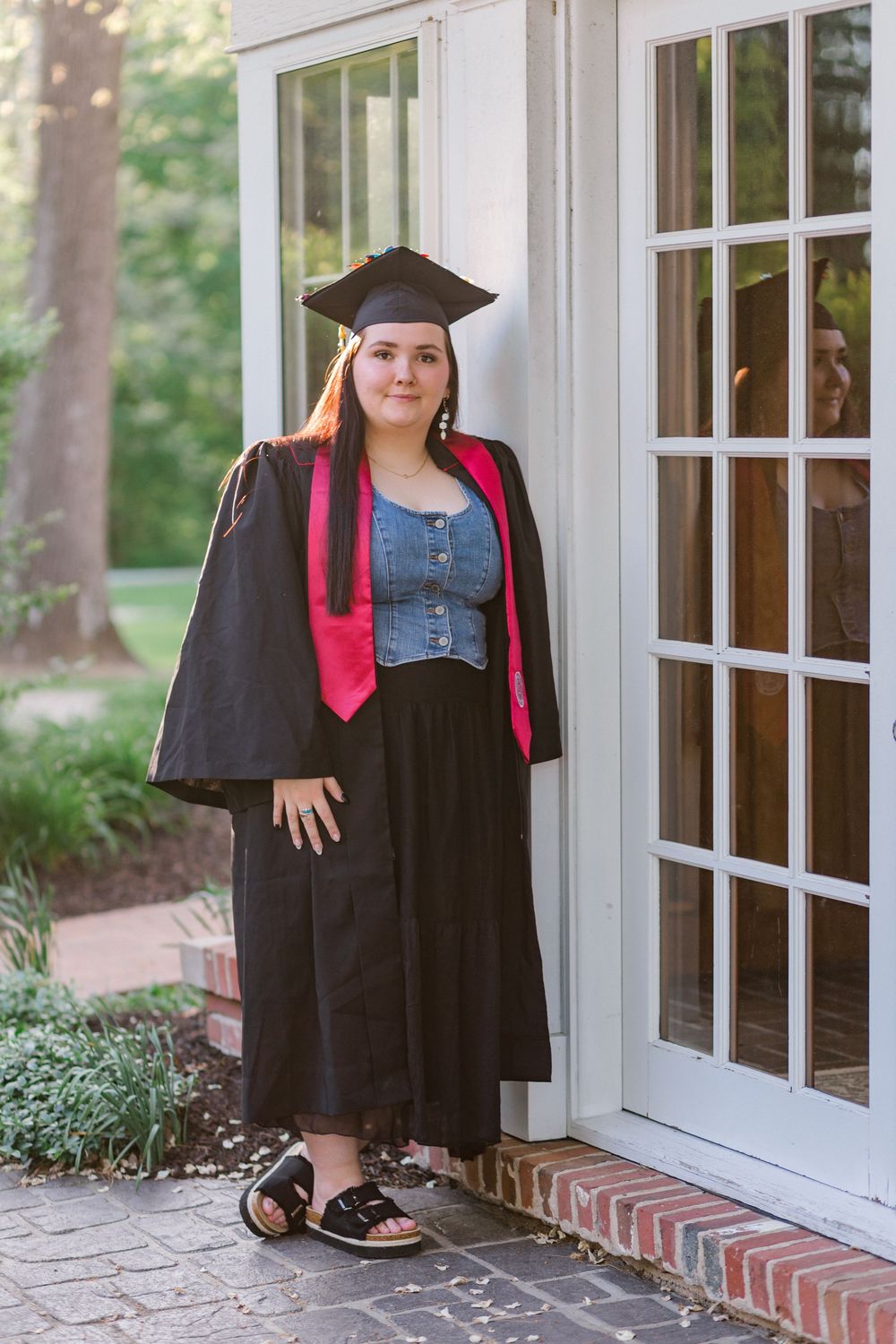 Young woman in black graduation cap and gown with pink stole poses proudly outside a white French door.