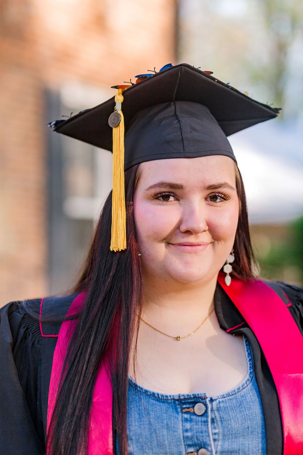 Young woman smiling in black graduation cap with yellow tassel and pink academic hood over denim top.