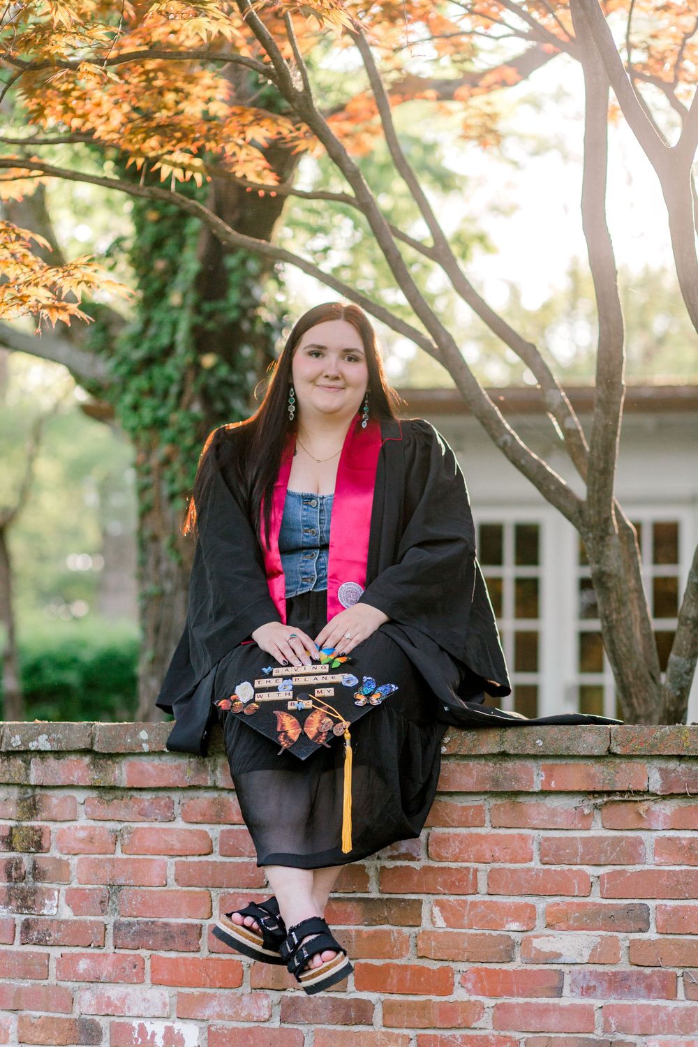 Graduate in black robe with pink sash sits on brick wall holding decorated cap outdoors.