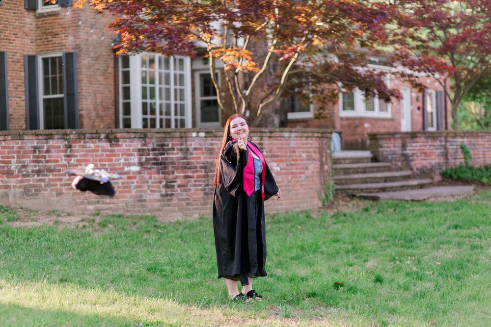 Young woman in black graduation gown with pink stole posing outdoors near brick building and red maple tree.