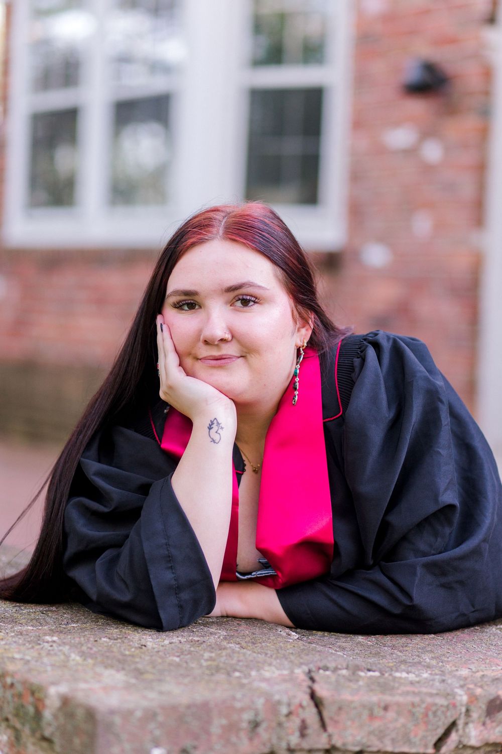 Graduate in black robe with pink stole poses on stone ledge near brick building.