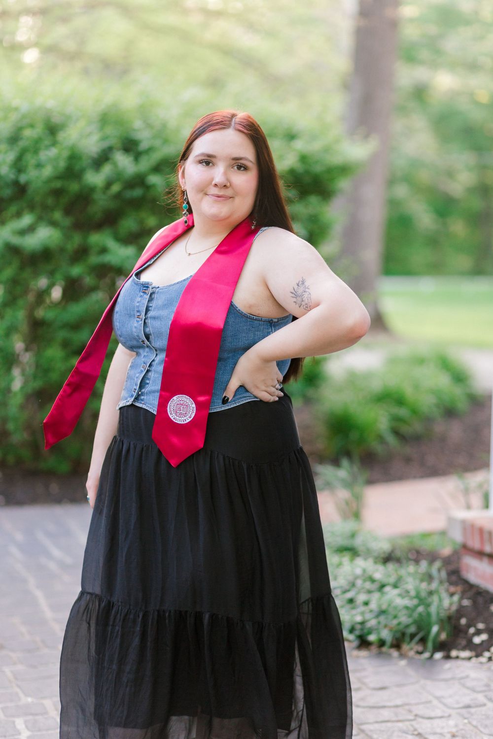 Graduate wearing denim top with red academic stole poses on outdoor brick pathway.