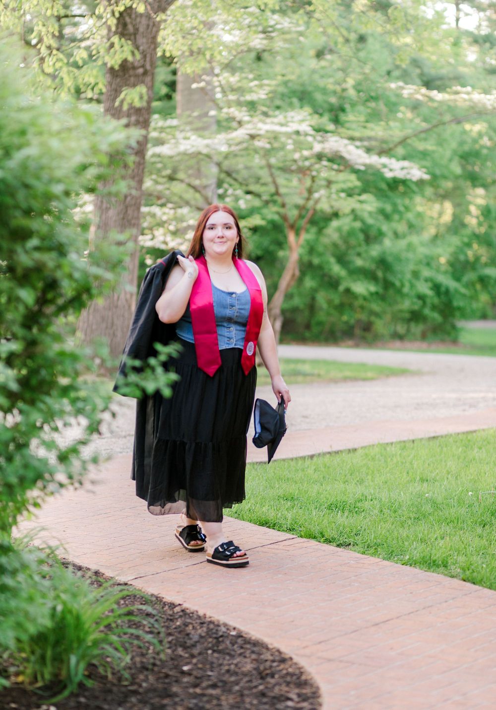 A smiling graduate walks along a tree-lined path carrying her black cap and gown with a pink graduation stole.