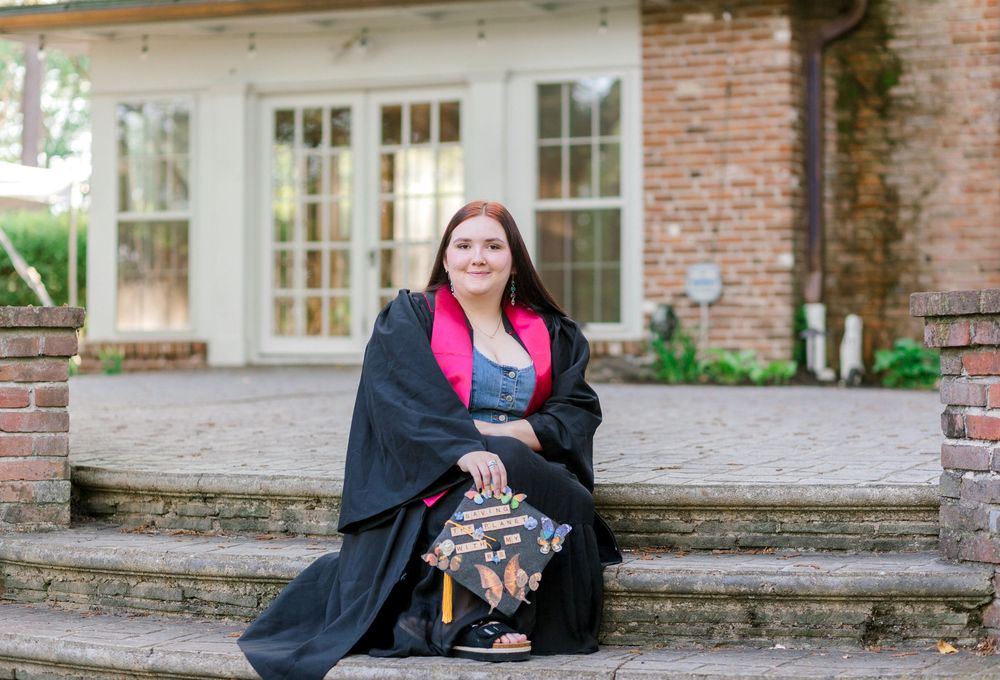 Young woman in graduation gown and pink sash sitting on brick steps outside a house.