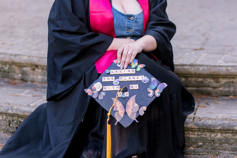 Graduate in black gown with decorated Scrabble tile mortarboard cap and yellow tassel at outdoor ceremony.