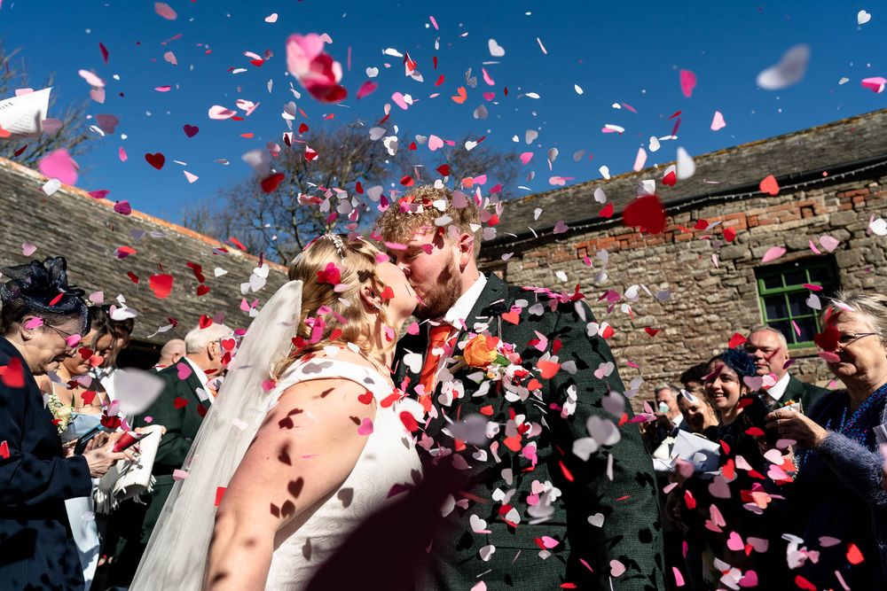 Bride and Groom share a kiss whilst confetti being thrown photographed by Devon Wedding photographer at Ash Barton Estate.