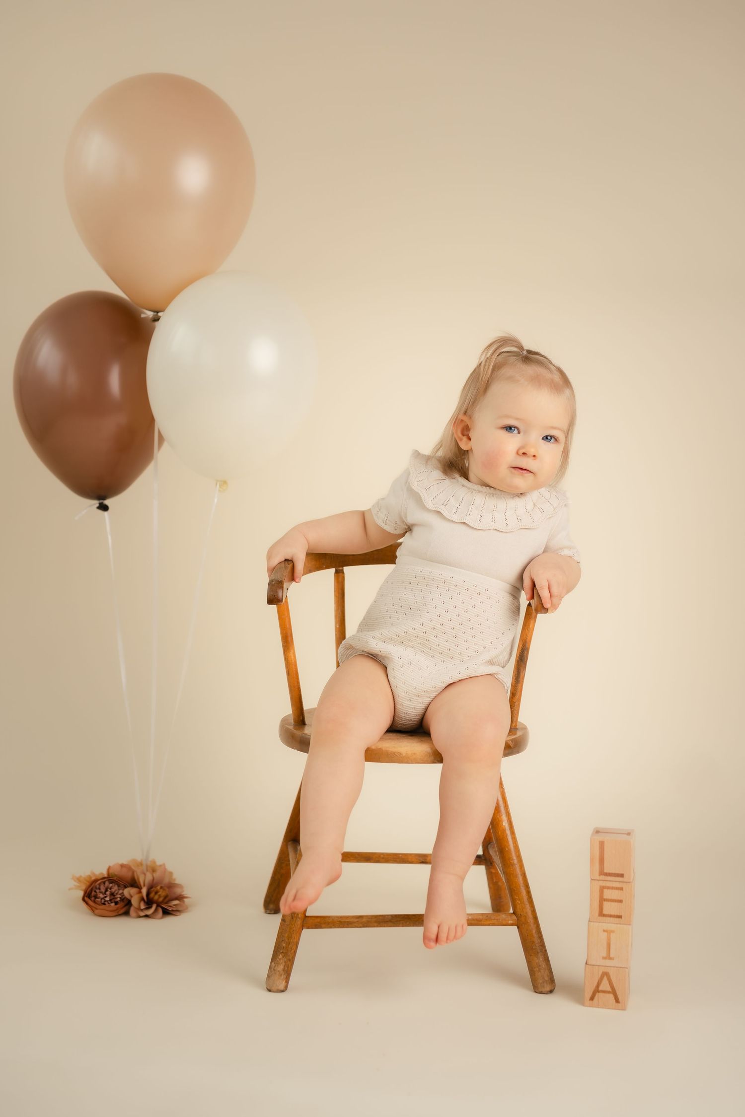 Baby girl named Leia sits on wooden chair with beige and brown balloons celebrating her first birthday.