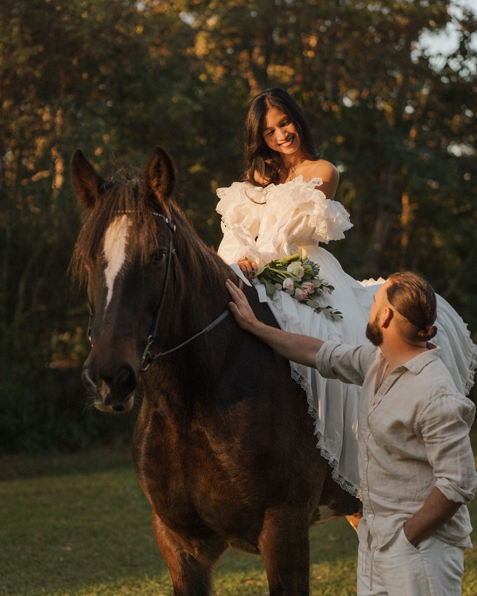 Bride in white gown sits on dark horse while groom stands beside her in golden hour light outdoors photoshoot with a horse in sydney