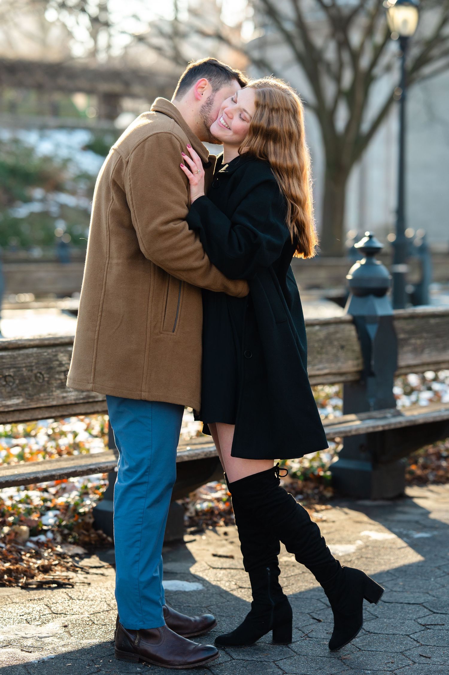 Couple embracing and smiling outdoors in winter coats, woman in black dress and knee-high boots, man in tan jacket.