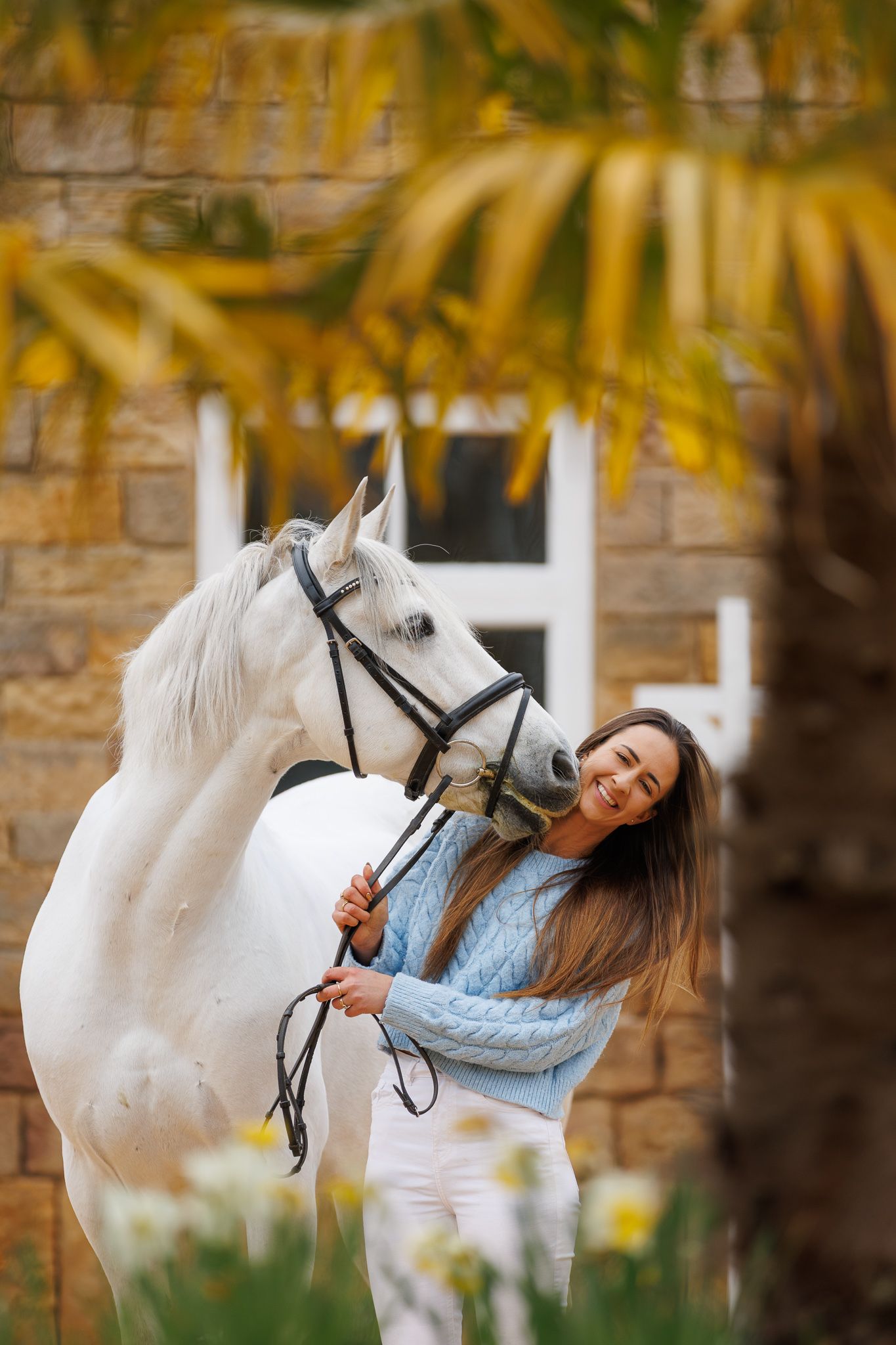 A beautiful bay horse wearing a black bridle stands next to someone in a white lace dress in an outdoor setting.