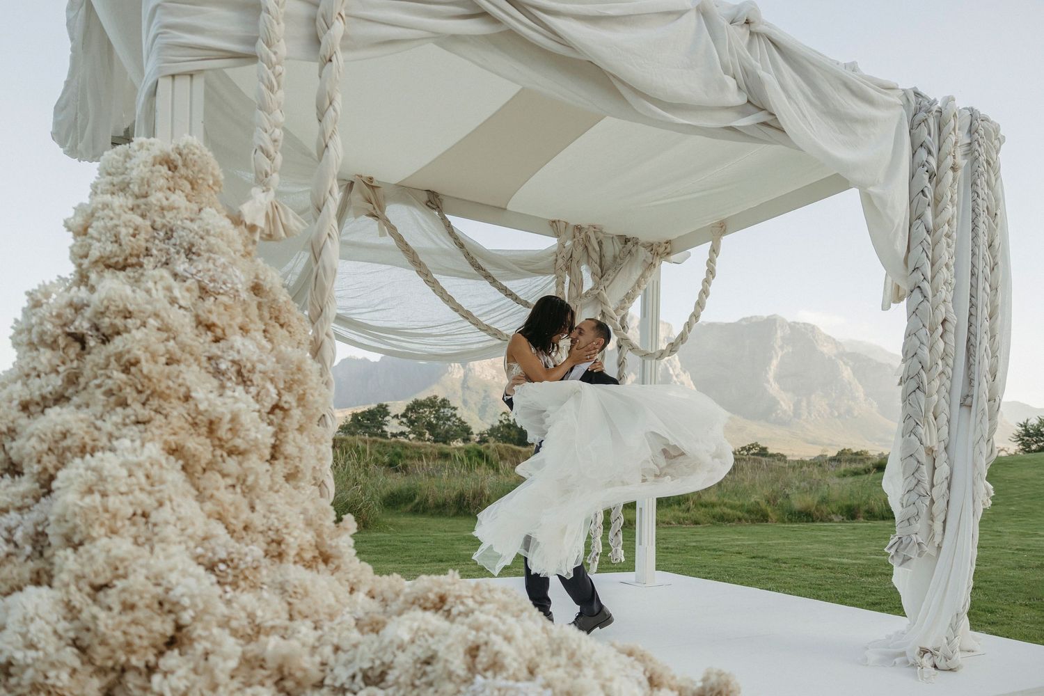 Bride and groom share romantic dip kiss under elegant white canopy with lush floral arrangements at outdoor wedding ceremony.