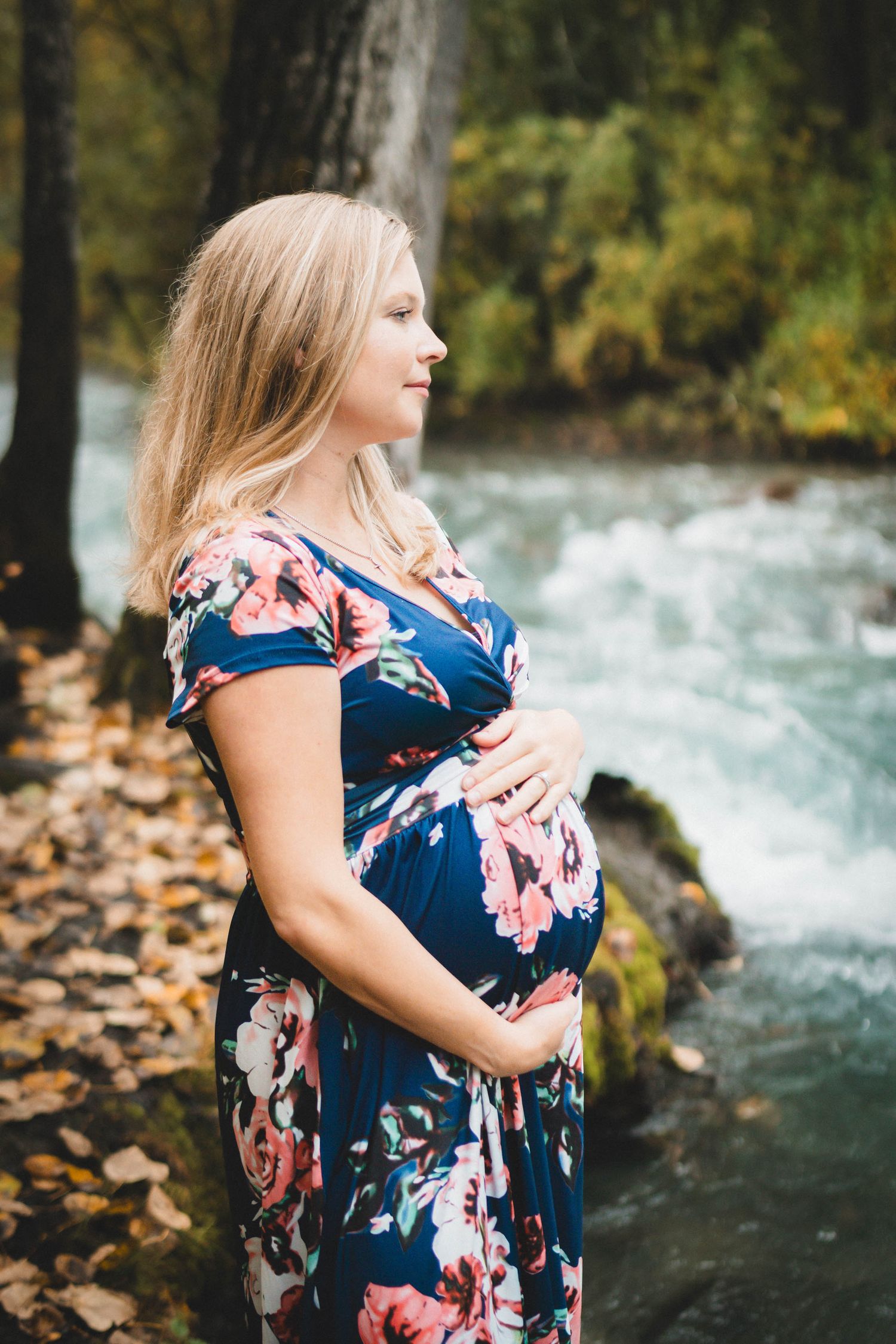 Pregnant woman in floral dress cradling her baby bump beside a flowing river in an autumn forest setting.