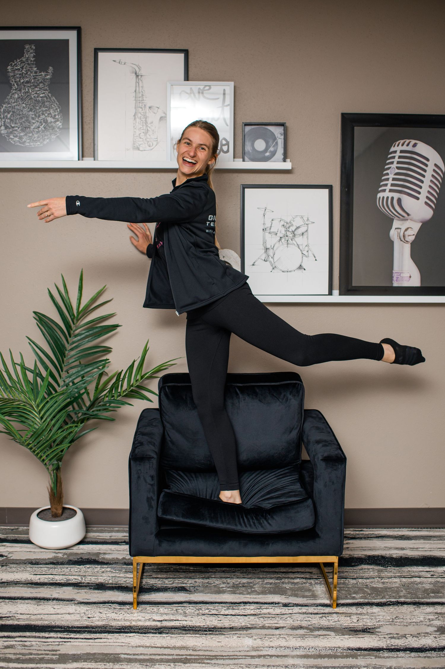 Woman in black outfit balancing on one leg atop a velvet chair, posing playfully in a music-themed room.