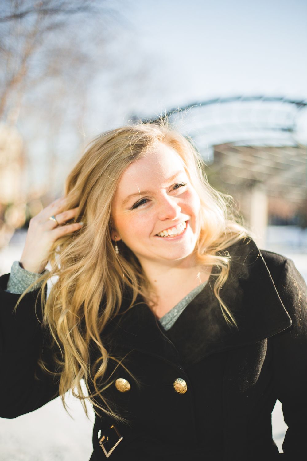 Smiling young woman with blonde wavy hair wearing a black coat outdoors on a sunny winter day.