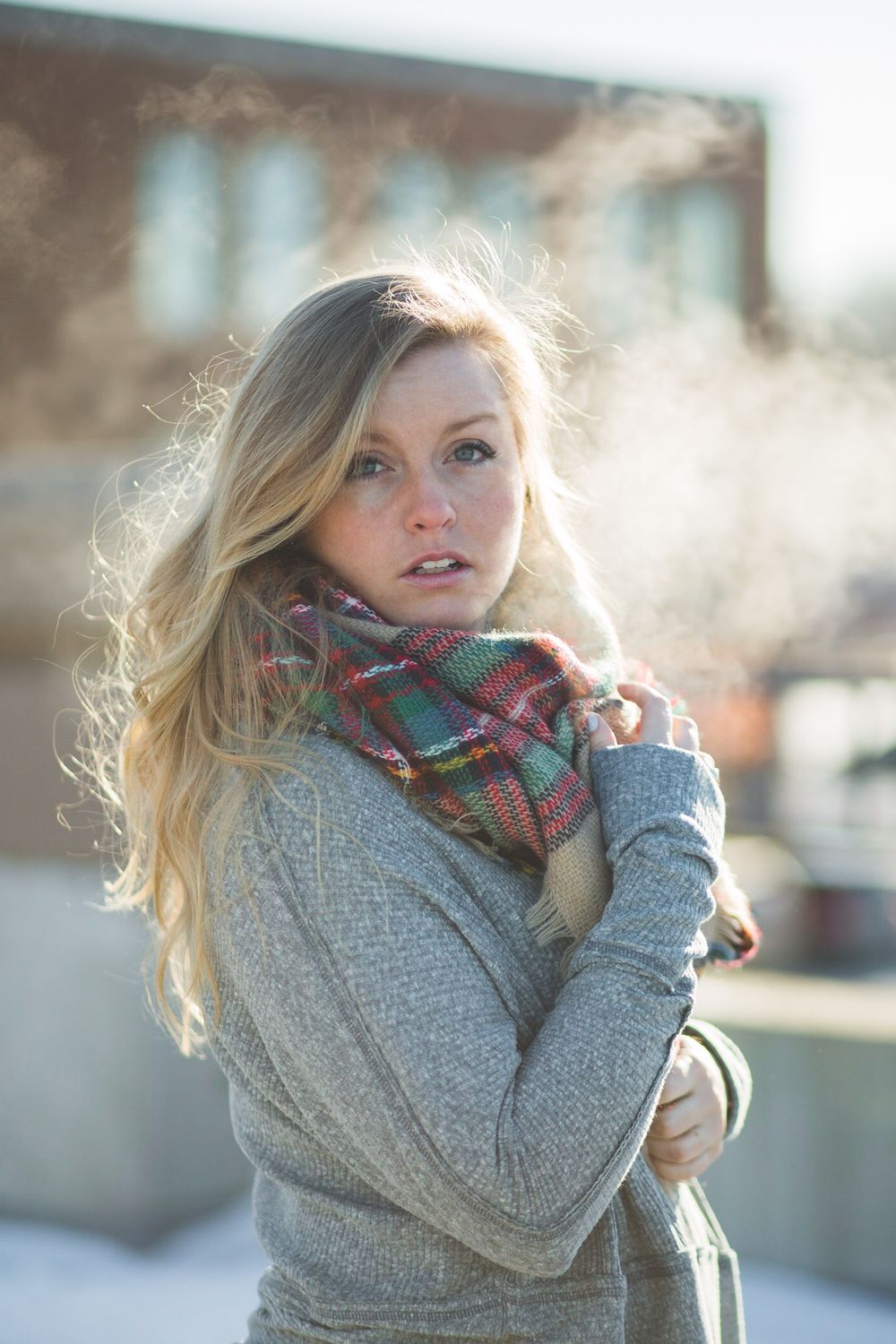 Young blonde woman in gray sweater and plaid scarf hugging herself outdoors in winter sunlight.