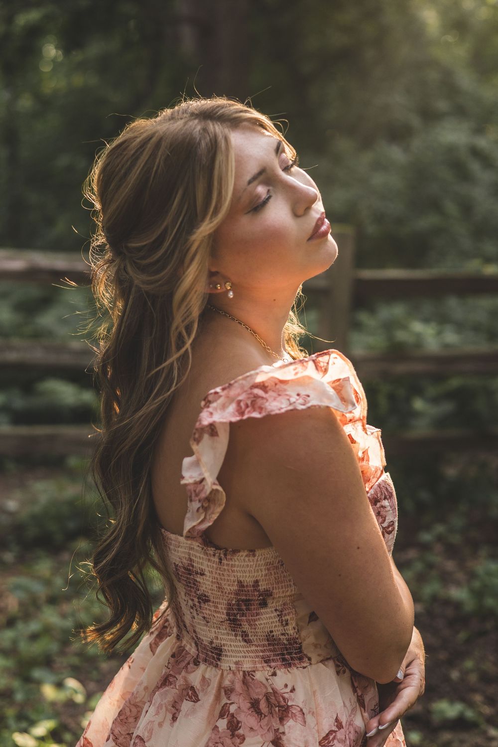Young woman in floral off-shoulder dress poses gracefully outdoors in golden sunlight with lush greenery background.