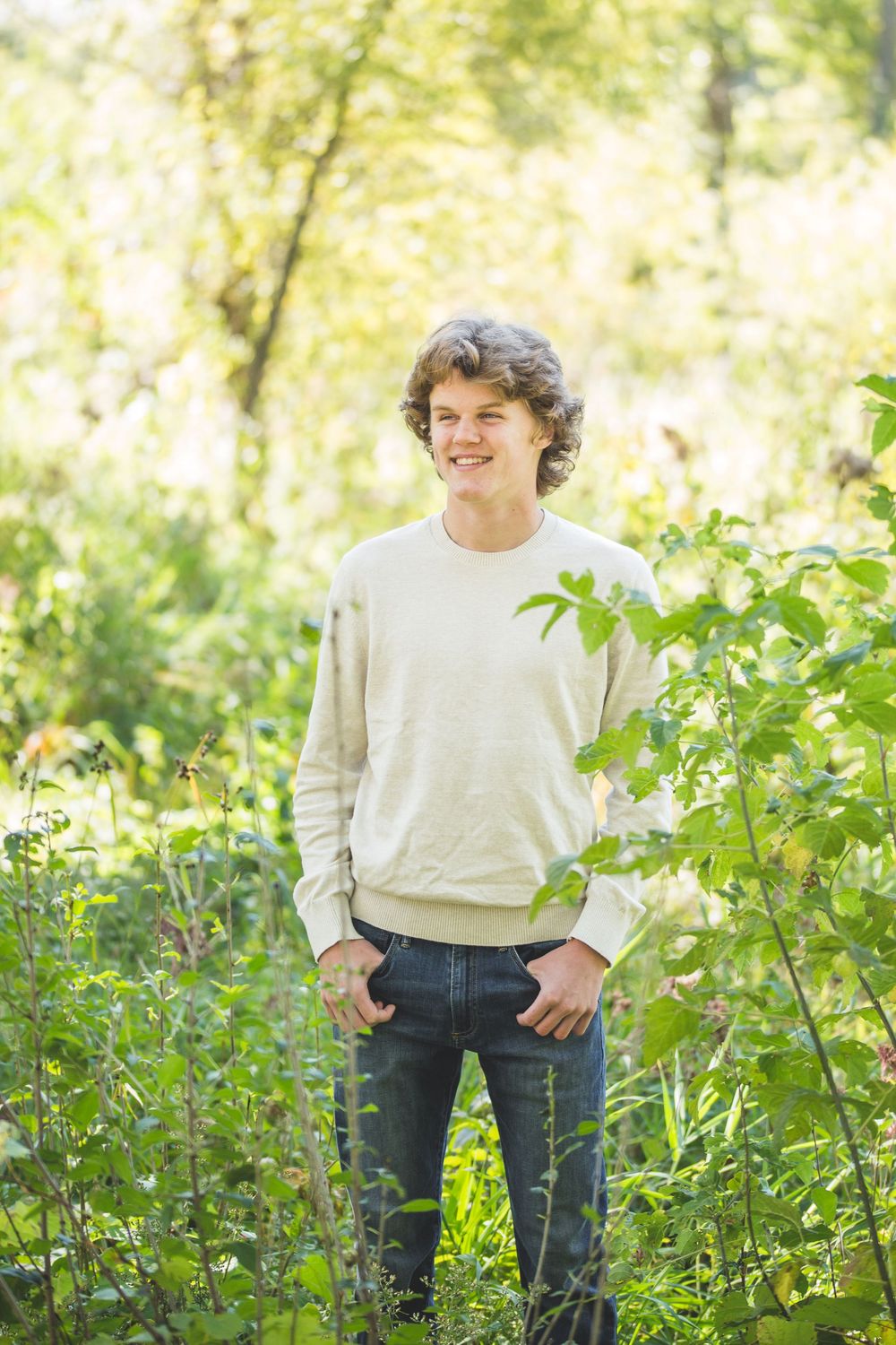 Smiling young man in cream sweater and jeans standing outdoors among lush green plants and trees.