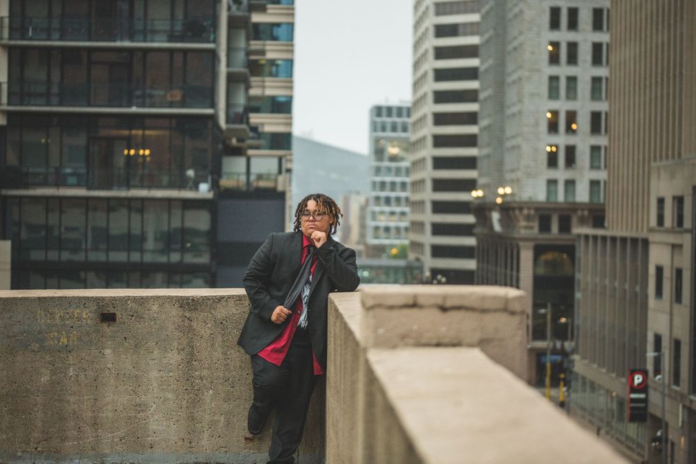 Person in black jacket and red shirt talking on phone on urban rooftop with city skyscrapers in background.