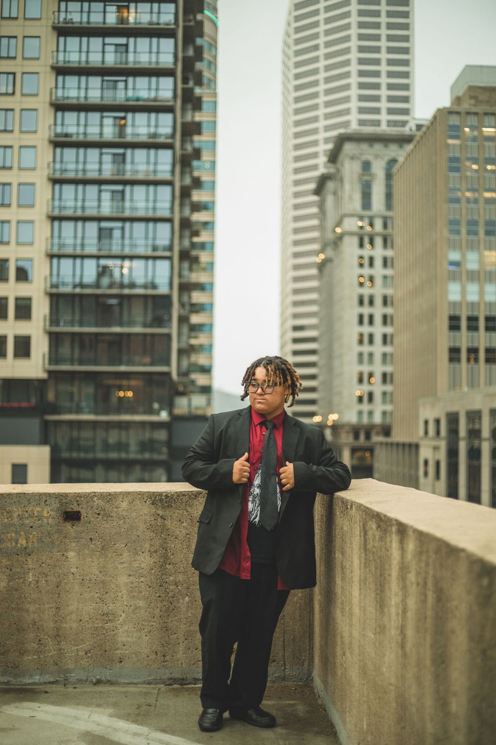 Young man in black suit and red shirt stands on urban rooftop with city skyscrapers in background.