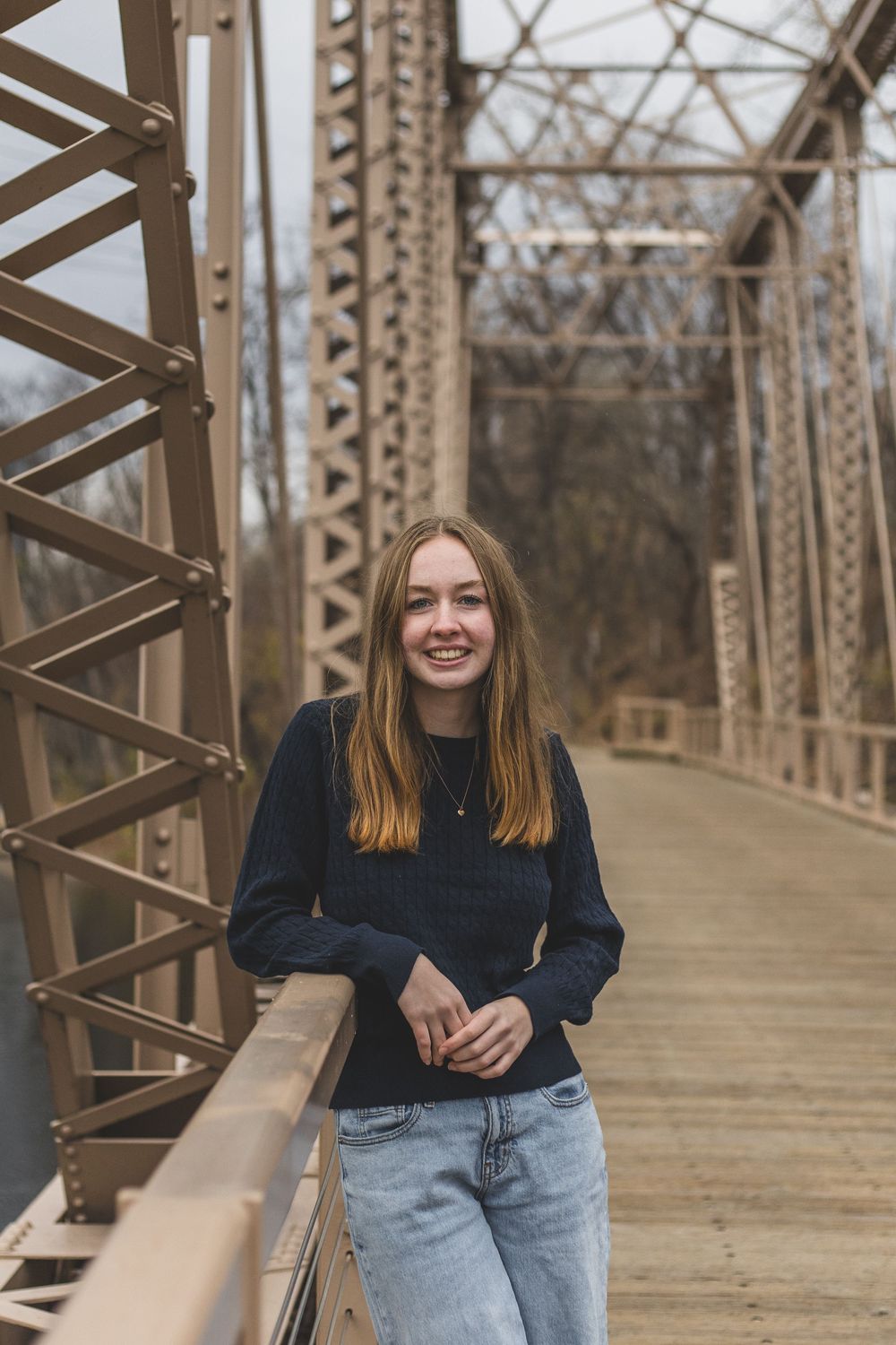 Smiling young woman in dark sweater and jeans posing on a rustic iron truss bridge.