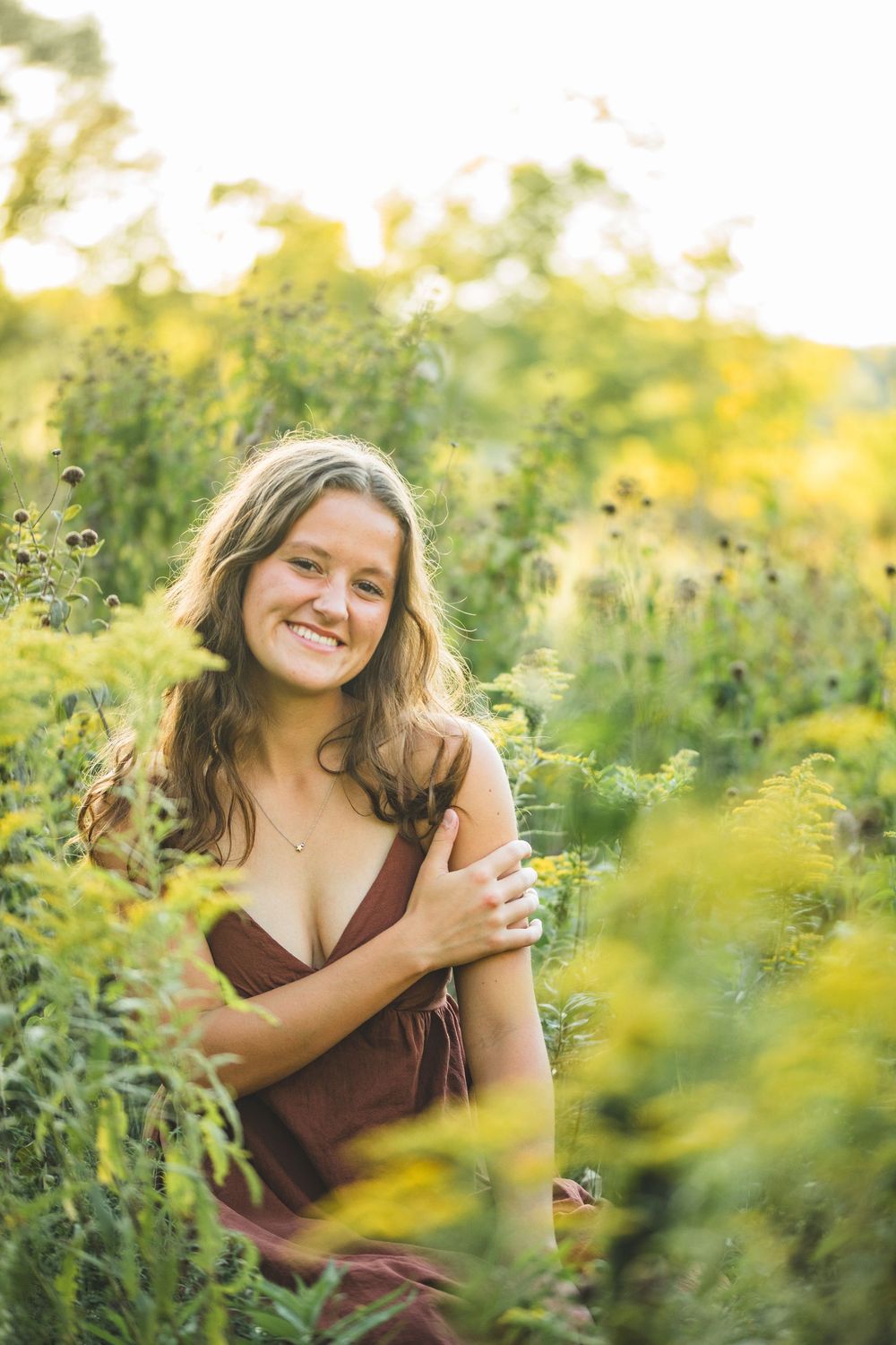 Happy young woman in brown dress smiles warmly while standing among vibrant wildflowers at sunset.