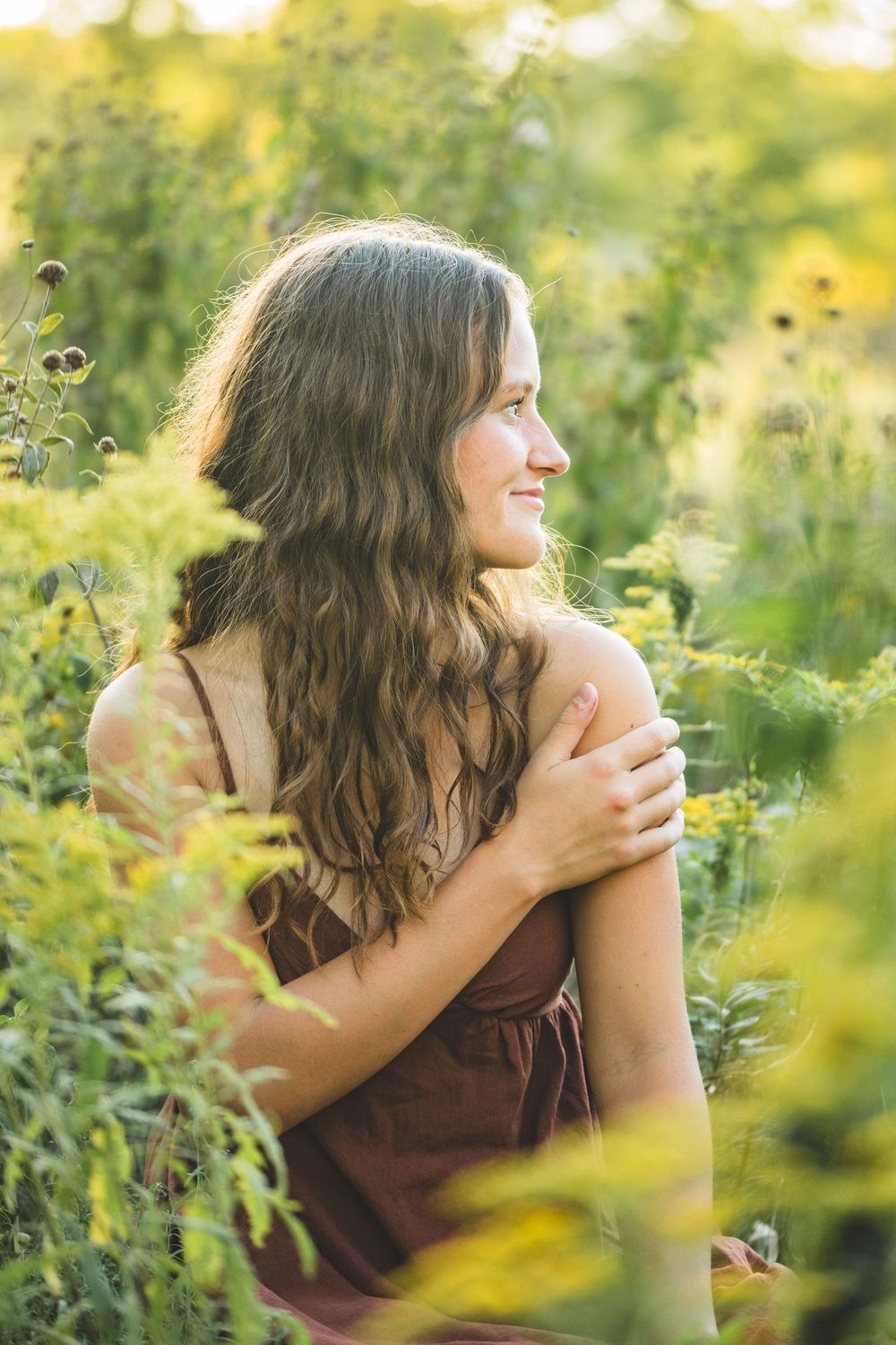Young woman with long wavy hair in a brown dress, sitting among lush green wildflowers in golden sunlight.