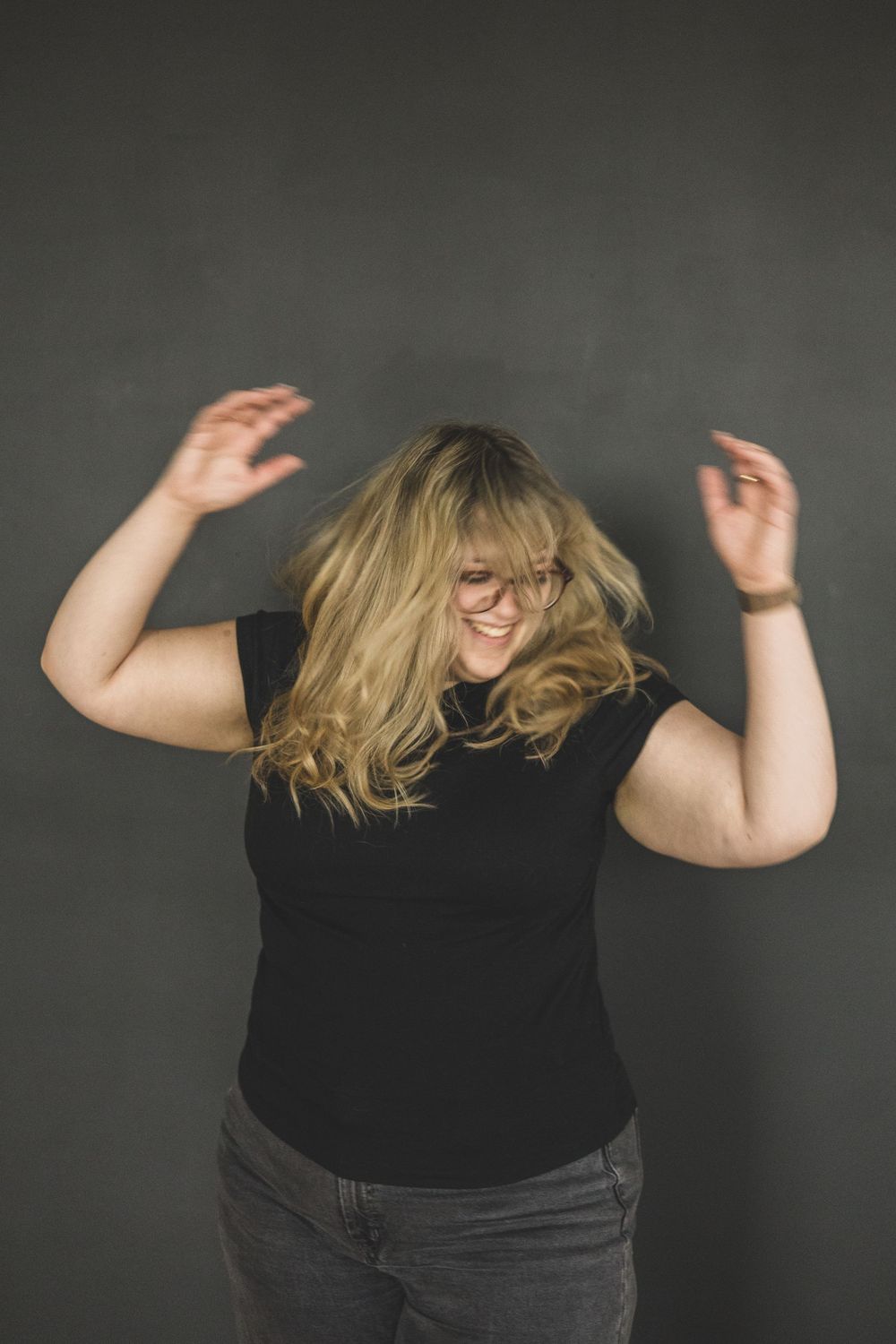 Joyful blonde woman in black t-shirt dancing with hands raised against gray background.