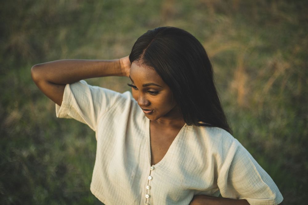 Young Black woman in white top poses outdoors against green grass background, hand raised to head.