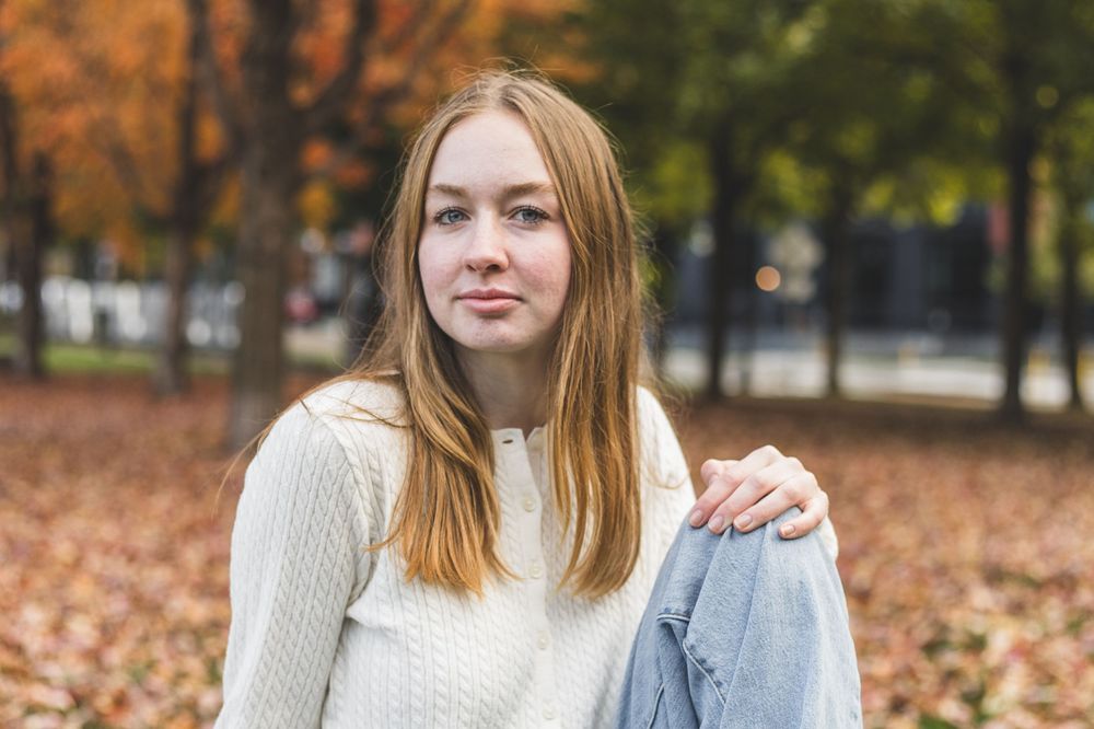 Young woman with auburn hair in white sweater sitting in autumn park with colorful fall foliage background.