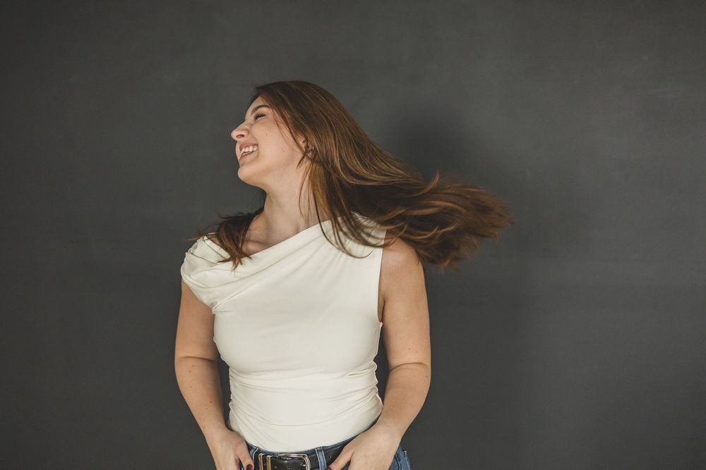 Young woman in white top tossing her brown hair, smiling against a dark gray background.