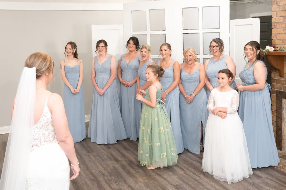 Bride reveals wedding look to bridesmaids in blue gowns, flower girl in green dress, in elegant venue.