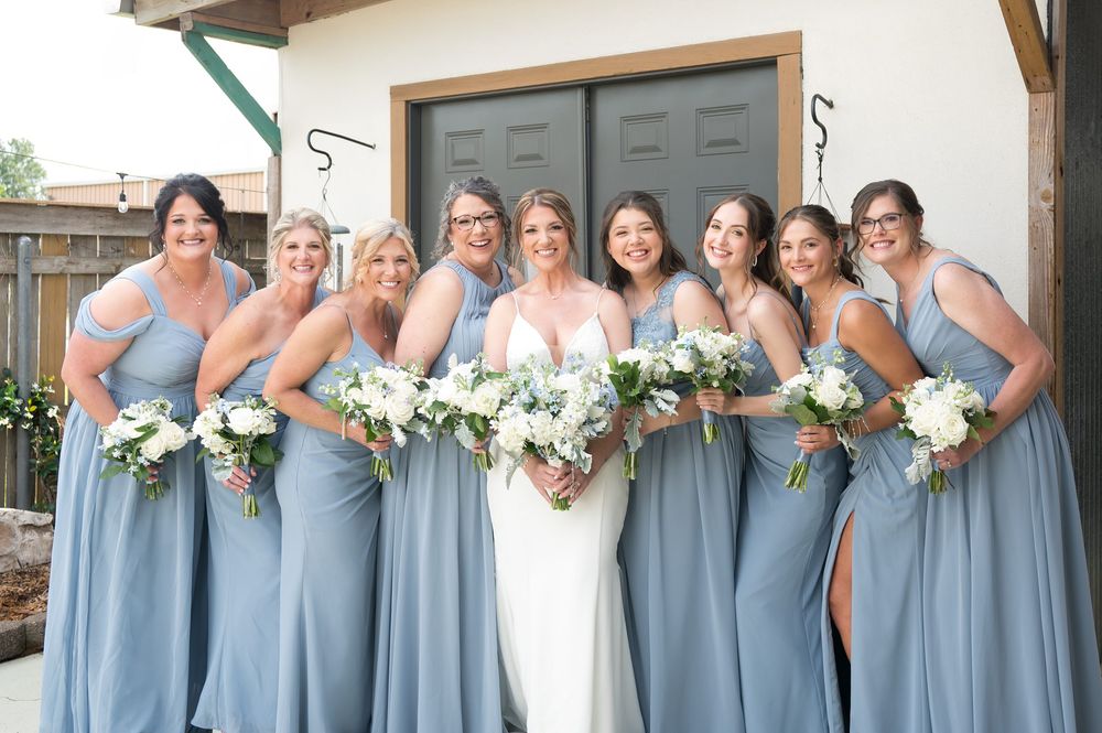 Bride in white gown with eight bridesmaids in dusty blue dresses holding white floral bouquets outdoors.