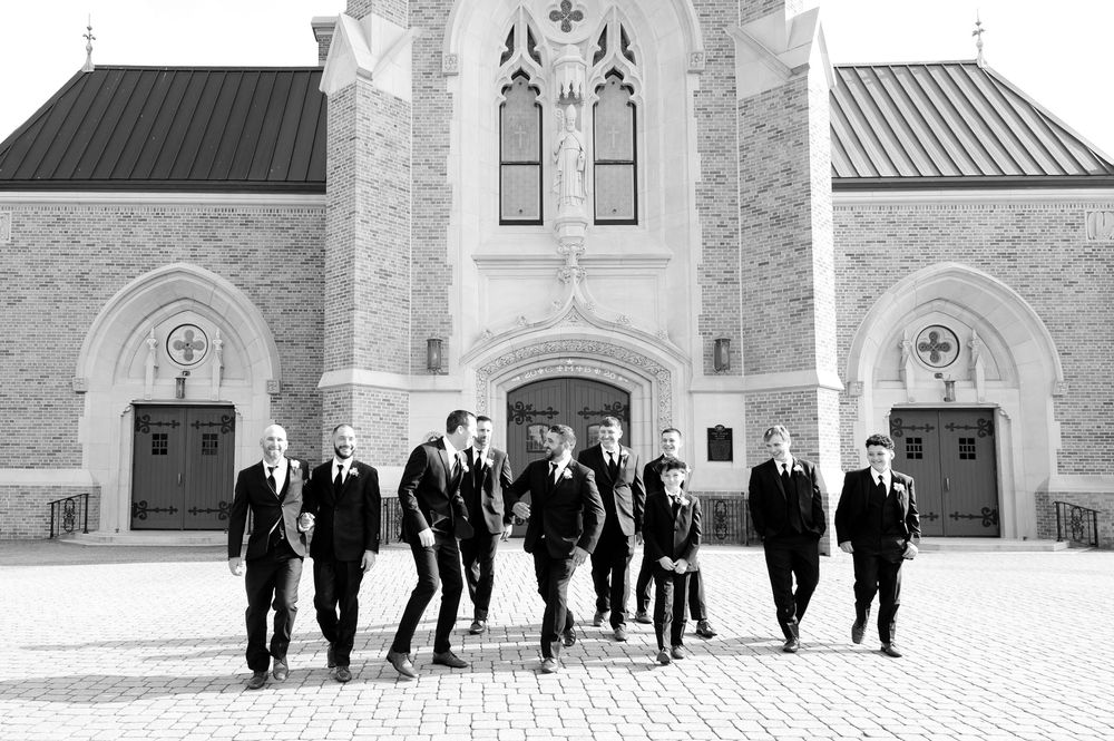 Groom and groomsmen in black suits walking together in front of a stone church in black and white photo.