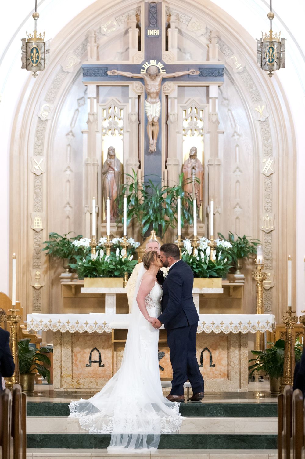 Bride and groom share first kiss at ornate Catholic church altar adorned with white flowers and crucifix.