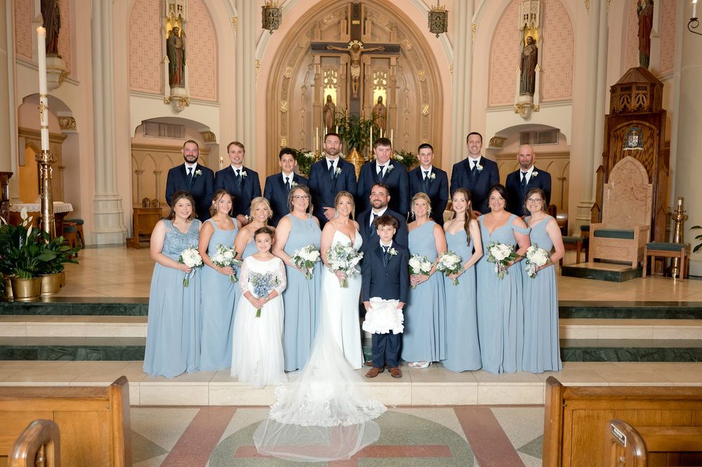 Wedding party in dusty blue dresses and navy suits poses with bride and groom inside a Catholic church.