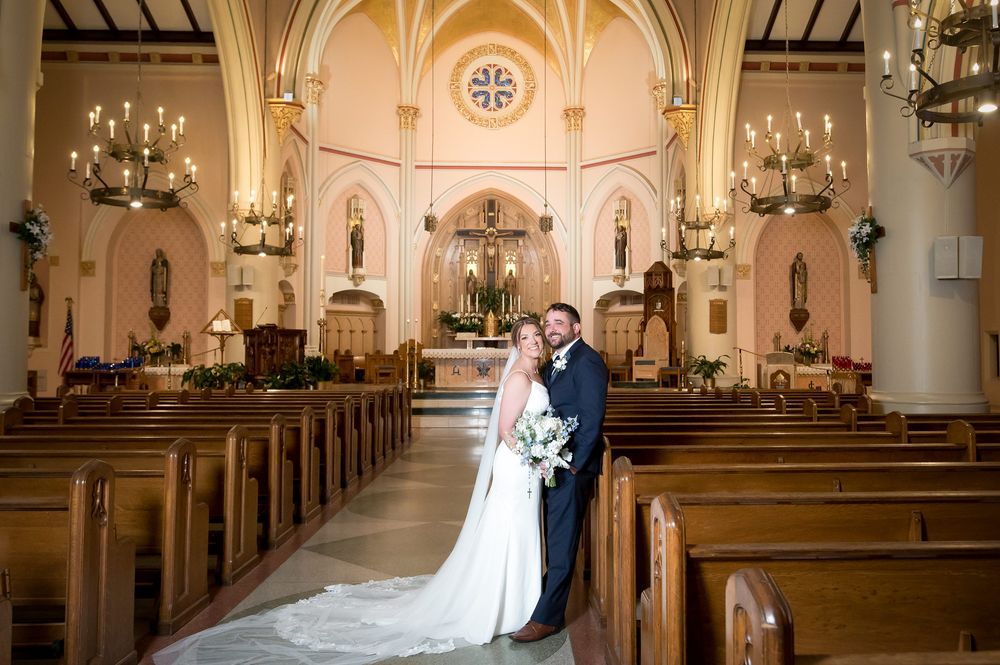 Bride and groom pose in elegant church aisle with wooden pews and chandelier after wedding ceremony.