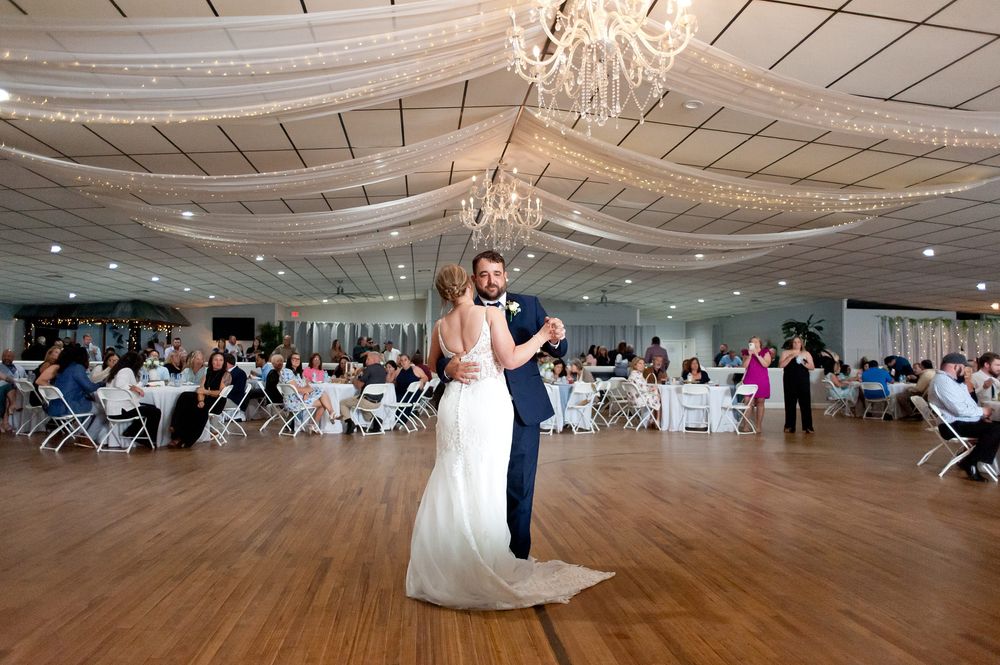 Bride and groom share first dance on wooden floor in elegant wedding reception venue with chandelier and guests seated.