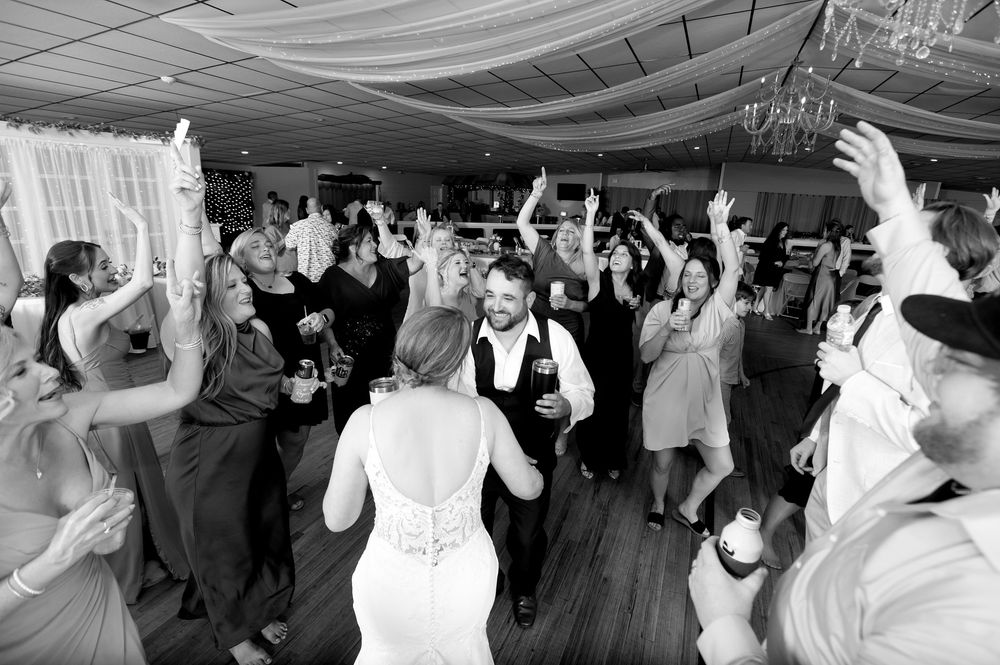 Black and white photo of bride and groom dancing with guests at a lively wedding reception celebration.