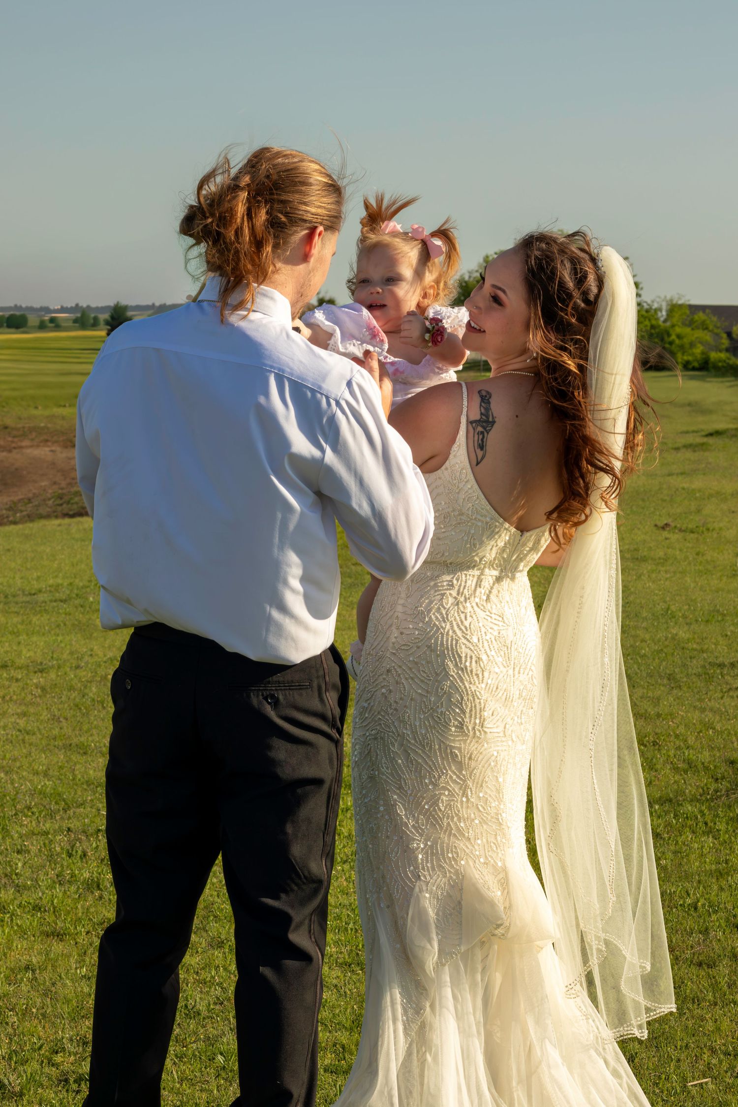 Bride and groom in wedding attire holding a baby girl outdoors on a green field on a sunny day.