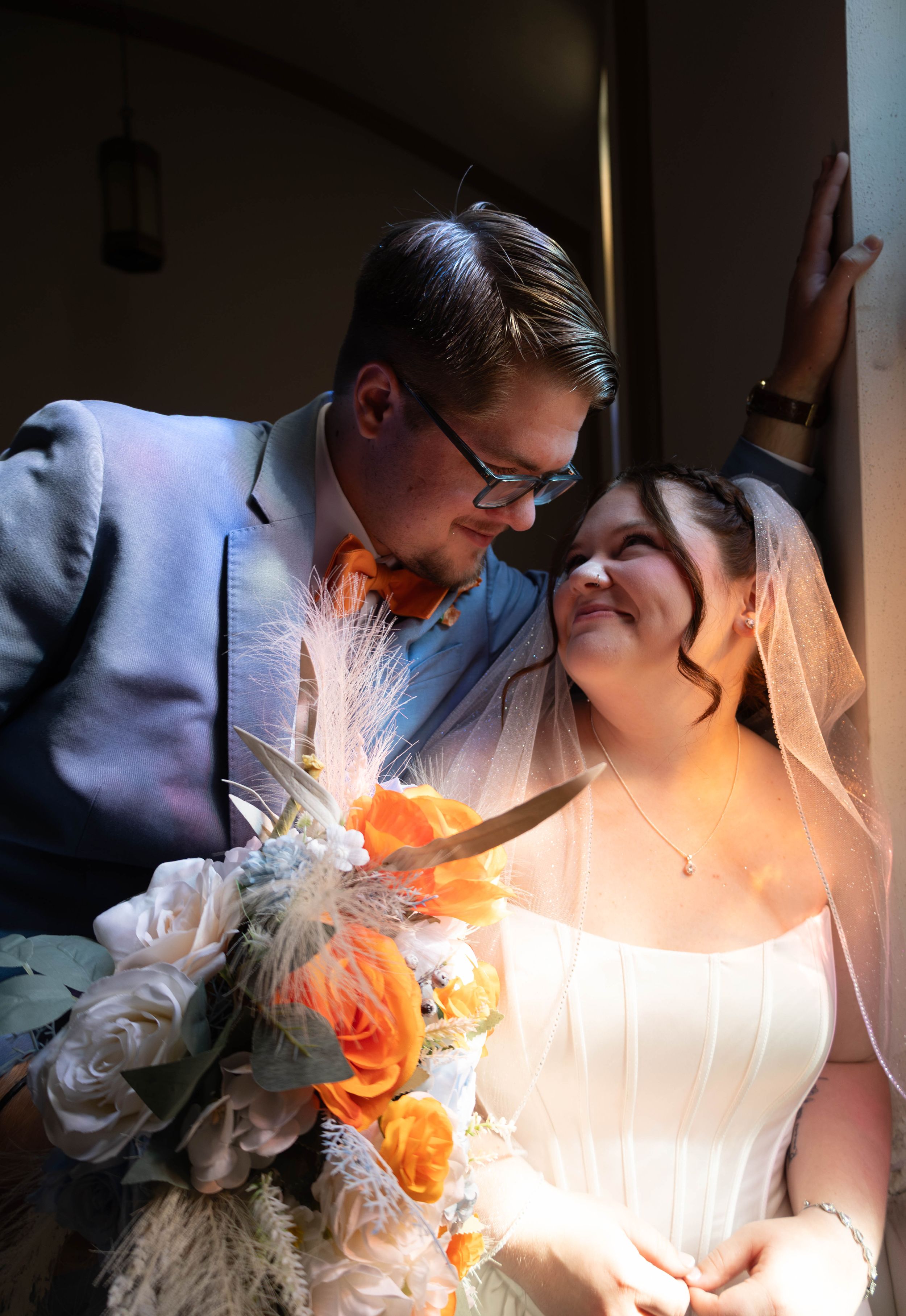 Bride and groom share romantic moment, bride holding orange and white bouquet, groom in blue suit with orange tie.