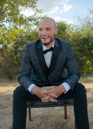 Smiling man in a dark floral tuxedo jacket with bow tie sits outdoors on a chair with trees in background.