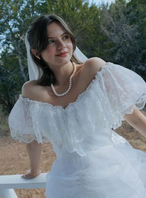 Young bride in white off-shoulder lace gown and pearl necklace with veil, smiling outdoors near white railing.